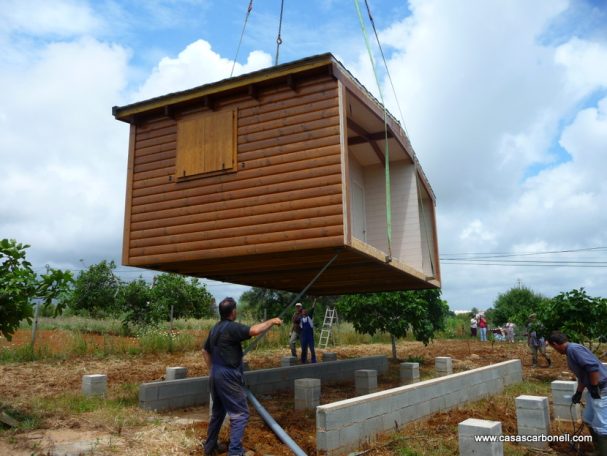 Base de muros corridos y pilares sobre riostra de hormigón soterrada Una casa de madera es levantada por grúas sobre una base en un terreno abierto.