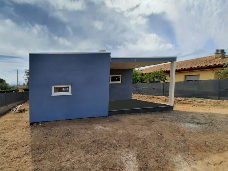 Casa moderna de color azul, con un porche y terreno seco alrededor.