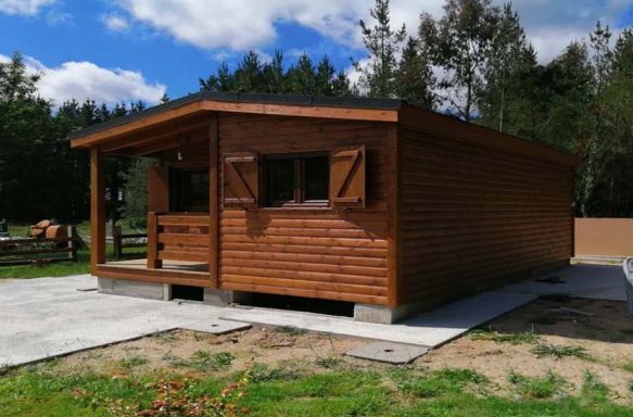 Casa prefabricada de madera marrón con ventanas y un porche, situada en un entorno natural.