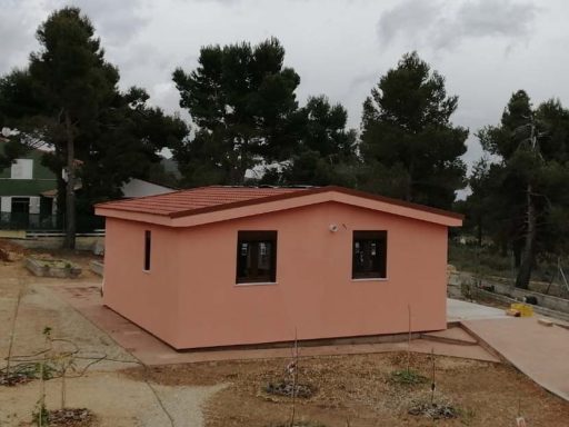 Casa prefabricada de color rosa con ventanas oscuras, situada en un paisaje rural.