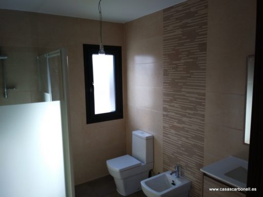 Baño moderno con lavabo, inodoro y ducha, paredes de azulejos claros y ventana.