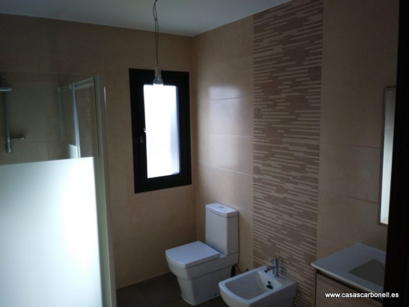 Baño moderno con lavabo, inodoro y ducha, paredes de azulejos claros y ventana.