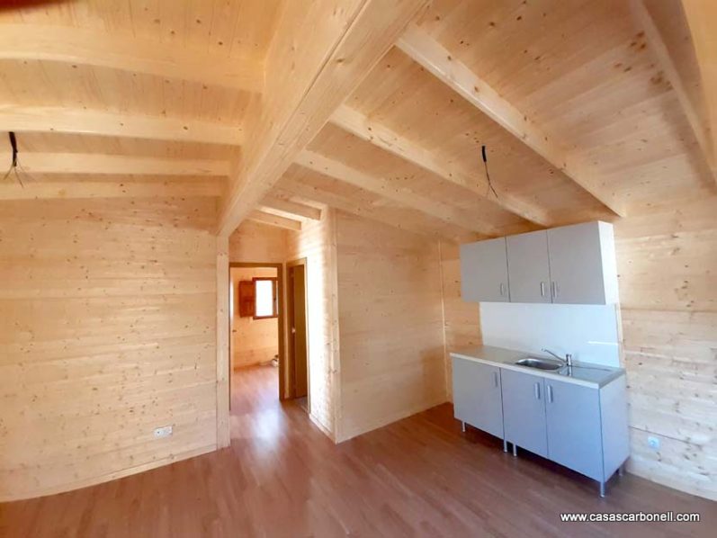 Interior de una casa de madera con techo alto y cocina pequeña. Suelo de madera.