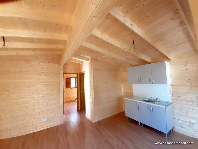 Interior de una casa de madera con cocina y ventanas iluminadas. Suelo de madera clara.