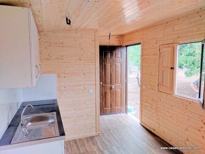 Interior de una pequeña cabaña de madera con cocina y puerta de acceso.