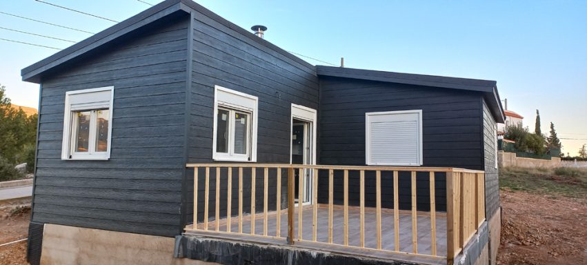 Casa prefabricada moderna de paredes negras y porche con barandilla de madera.