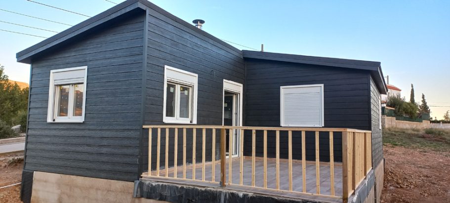Casa prefabricada de una planta con fachada negra y porche de madera en un entorno rural.