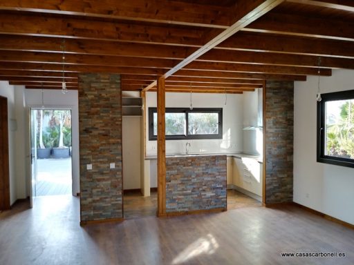 Interior de una sala con paredes de piedra y cocina integrada, luz natural y techos de madera.