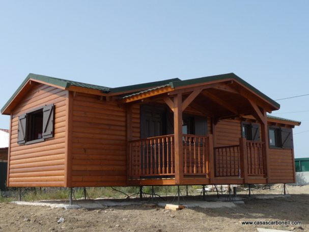 Casa prefabricada colocada sobre pies de apoyo metálicos regulables, sin cimentación Casa prefabricada de madera con porche, ventanas y un techo a dos aguas en un entorno despejado.