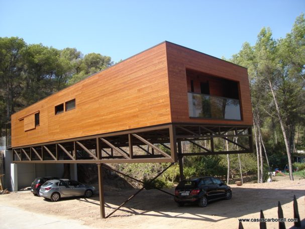Base metálica con vigas de celosía elevada Casa moderna de madera elevada sobre pilotes, rodeada de árboles y coches estacionados.