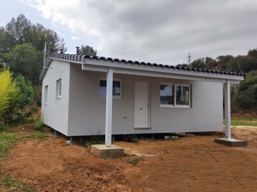 Casa prefabricada pequeña de un solo piso con paredes blancas y techo a dos aguas, rodeada de naturaleza.