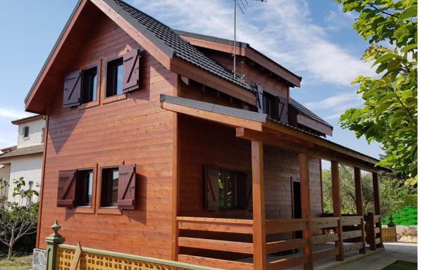 Casa prefabricada de madera con balcón, ventanas con persianas y entorno natural verde.