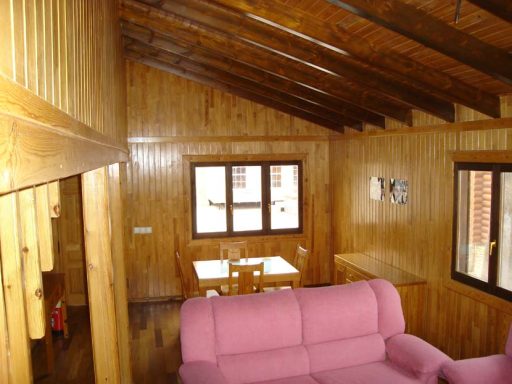 Interior de una cabaña de madera con sofá rosa y mesa de comedor. Ventanas iluminan el espacio.