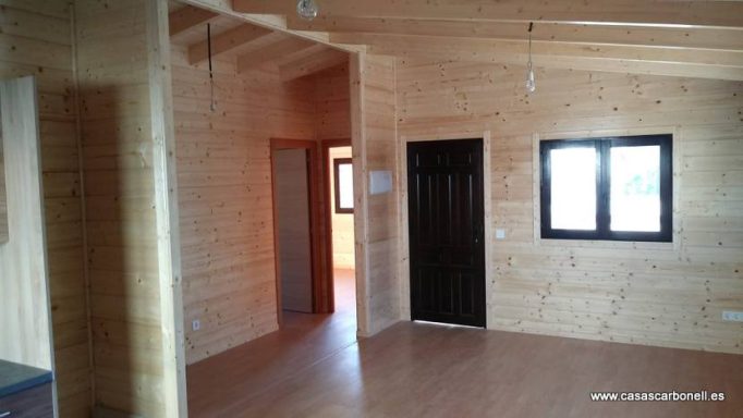 Interior de una casa de madera con paredes claras y puertas oscuras, iluminado por ventanas.