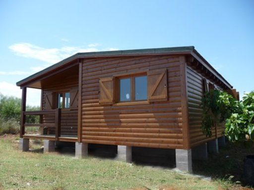 Casa prefabricada de madera de un solo piso con porche y ventanas con persianas, en terreno despejado.