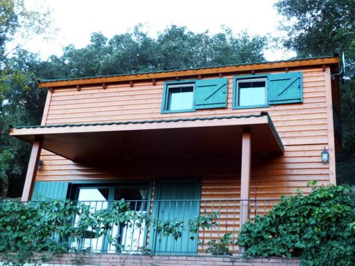 casa prefabricada de madera de dos plantas con ventanas verdes y porche frontal. Rodeada de vegetación.