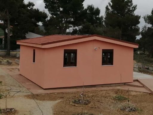 Casa prefabricada de color rosa con techo a dos aguas, rodeada de terreno y árboles.