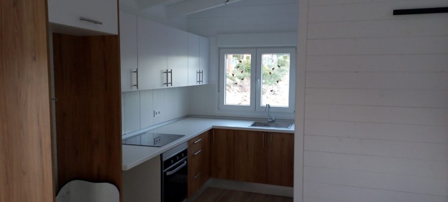 Cocina moderna con muebles de madera clara y encimera blanca, con ventanas iluminadas.