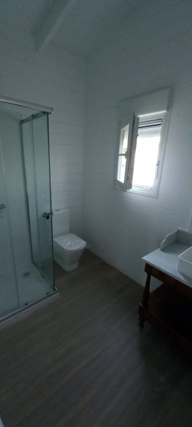 Baño moderno con ducha, inodoro, lavabo y ventana. Suelo de madera.