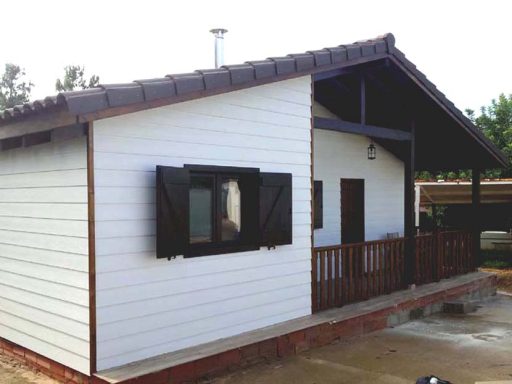 Casa prefabricada de un piso con tejado a dos aguas, paredes blancas y persianas de madera.