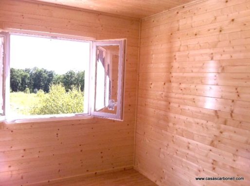 Interior de una habitación de madera con una ventana abierta que da a un paisaje verde.