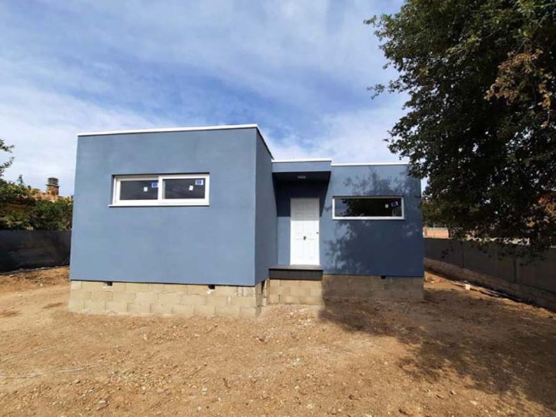 Casa moderna de color azul con ventanas y puerta blanca, ubicada en un terreno despejado.