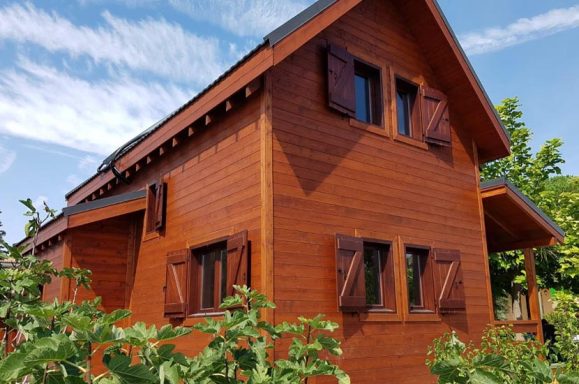 Casa prefabricada de madera de dos plantas con persianas, rodeada de vegetación y cielo despejado.