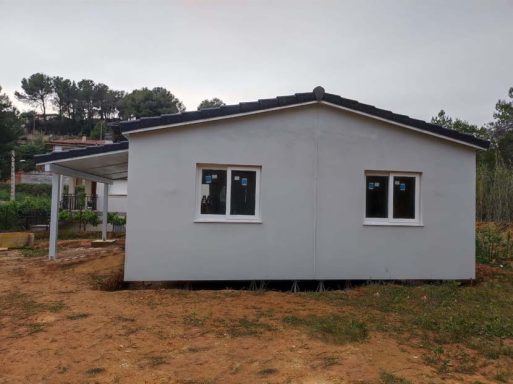 Casa prefabricada de un solo piso con paredes blancas y ventanas, rodeada de terreno al aire libre.