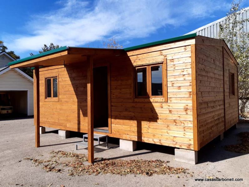 Casa de madera con techo verde, ventanas y porche, ubicada en un entorno exterior.