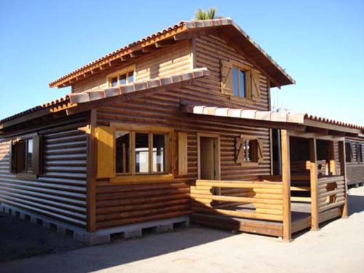 Casa prefabricada de madera de dos pisos con tejado a dos aguas y balcones en un entorno soleado.