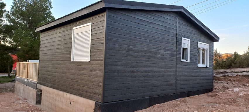 Casa prefabricada de madera oscura con ventanas blancas, situada en un entorno al aire libre.