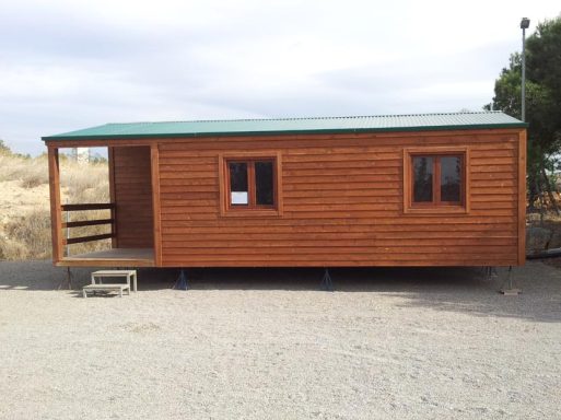 Casa de madera con ventanas, puerta frontal y tejado verde, ubicada en un entorno árido.