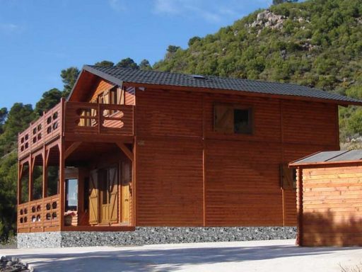 Casa prefabricada de madera de dos pisos con balcón, rodeada de árboles y un paisaje montañoso.