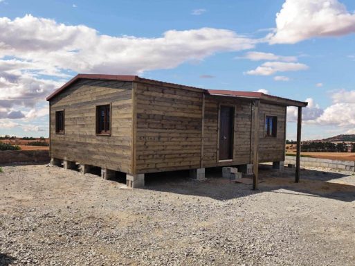 Casa de madera con techo rojo, ubicada en un terreno graveloso. Cielo nublado.