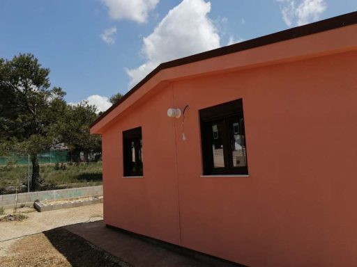 Casa prefabricada de color rosa con ventanas oscuras, situada en un área con árboles y cielo nublado.