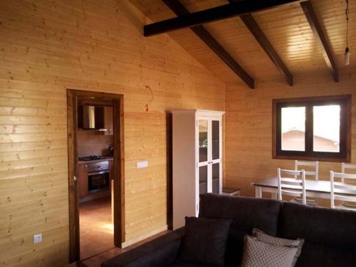 Interior de una cabaña de madera con una sala y una cocina visible. Mobiliario claro.