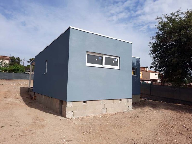 Casa moderna de un solo piso con paredes azules y ventanas, situada en un terreno despejado.