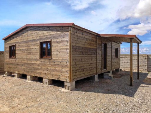 Casa de madera con ventanas, patio y base elevada, en un entorno despejado.