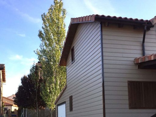 Casa prefabricada de dos plantas con revestimiento claro, árboles al fondo y cielo despejado.