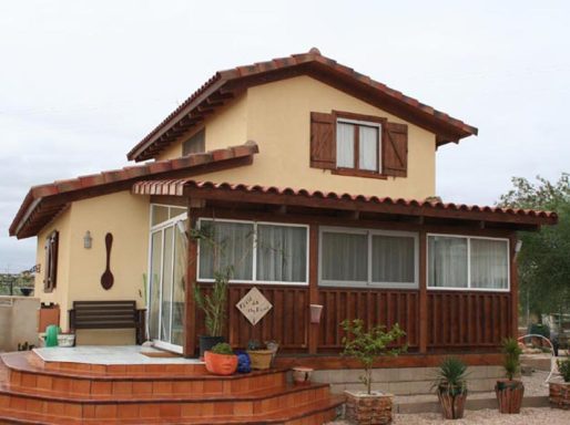 Casa prefabricada de dos plantas con paredes amarillas y detalles de madera, rodeada de plantas.