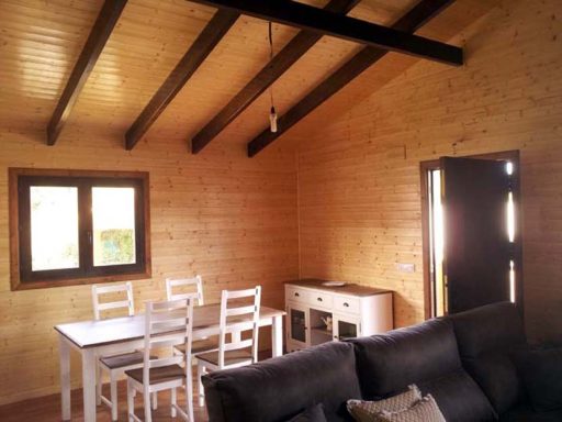 Interior de una cabaña de madera con mesa, sillas y ventana iluminada.