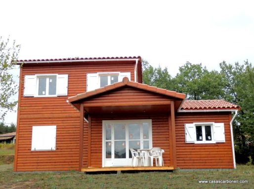 Casa prefabricada de madera de dos pisos con porche y ventanas blancas, rodeada de árboles.