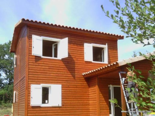 Casa prefabricada de dos plantas de madera con ventanas blancas y un trabajador en una escalera.