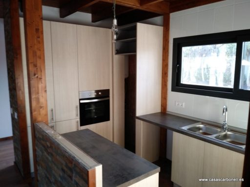 Cocina moderna con muebles de madera clara y encimera gris, ventana grande y electrodomésticos integrados.