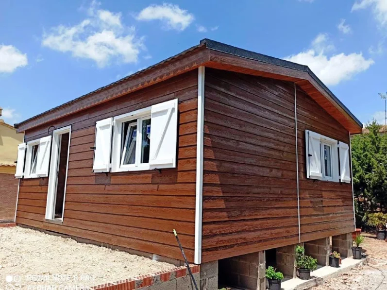 Casa prefabricada de madera con ventanas blancas, situada sobre pilotes en un paisaje despejado.