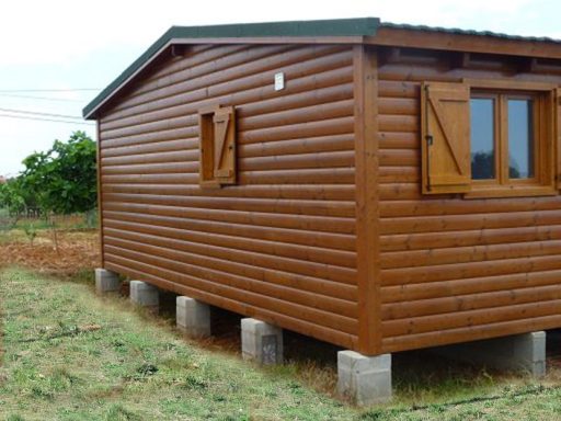 Casa prefabricada de madera con ventanas y contraventanas, situada sobre pilares de cemento en un campo.