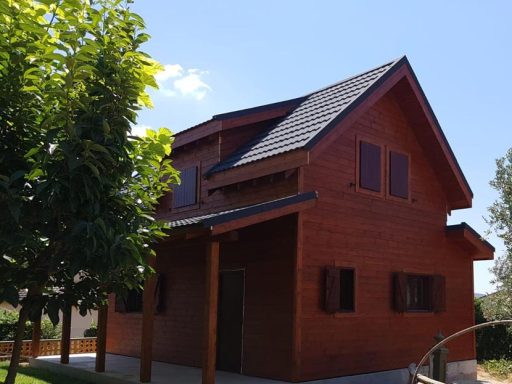 Casa prefabricada de madera de dos pisos con techo a dos aguas y ventanas grandes, rodeada de árboles.