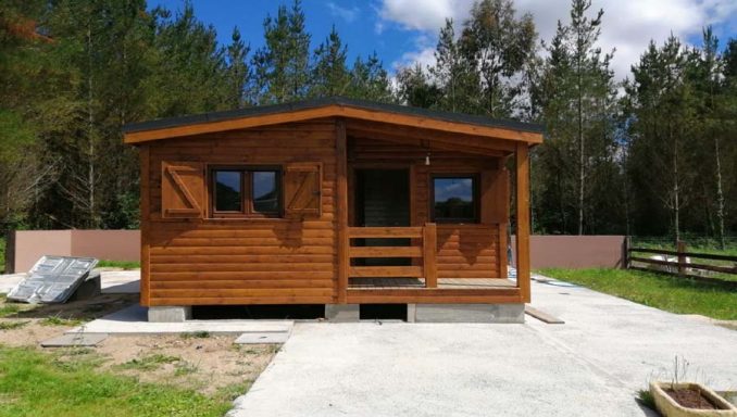 Casa prefabricada de madera con ventanas, rodeada de árboles y un camino de concreto.