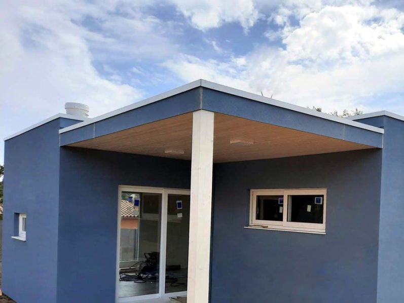 Casa moderna de color azul, con techo saliente y ventanas. Cielo nublado al fondo.