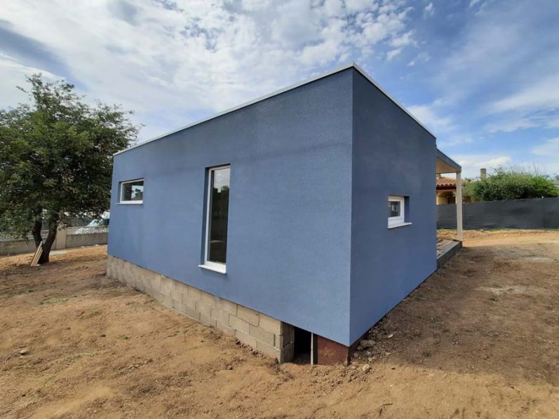 Casa moderna de color azul con ventanas, ubicada en un terreno seco y soleado.